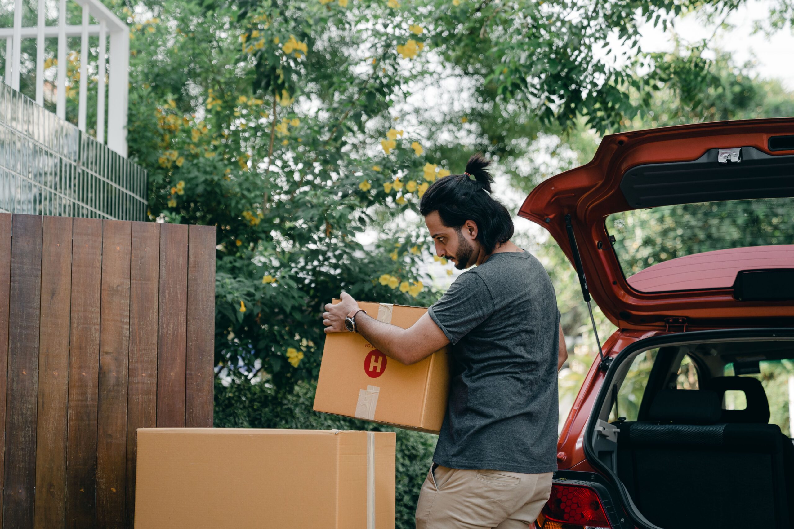 Side view of serious young ethnic man in casual clothes unloading cardboard boxes out of trunk of modern car in blooming green backyard while moving in new house